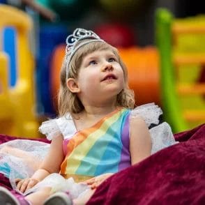 A little girl wearing a tiara sitting on a blanket at an early learning center.