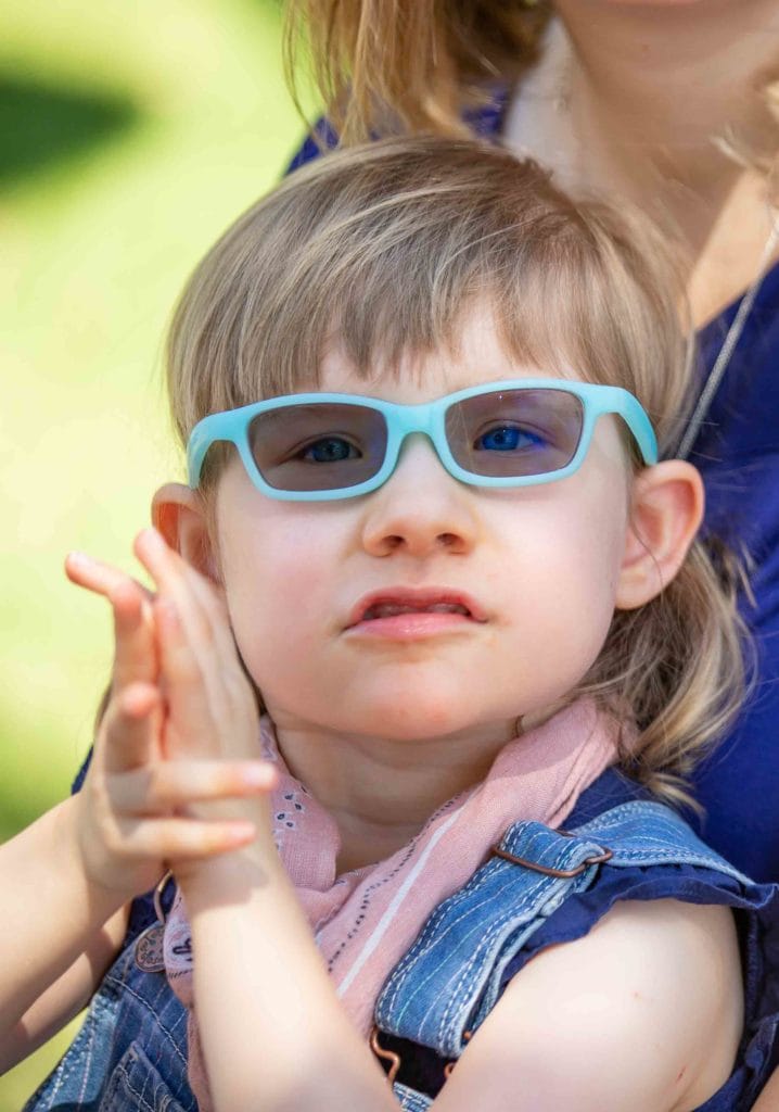A young girl with cerebral palsy, wearing glasses, clapping her hands at a pediatric therapy session.