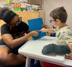 A woman in a mask provides pediatric therapy to a child at an early learning center.