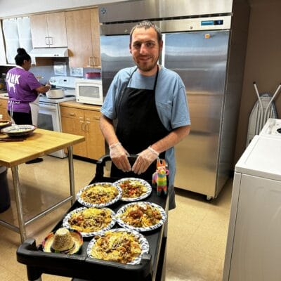 A man in an apron stands in a kitchen, smiling, next to a cart with several plates of food, as part of the adult day programs. Another person in a purple shirt is working diligently in the background.