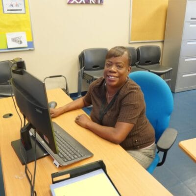 A woman sits at a desk with a computer in an office, smiling at the camera, as she researches adult day programs near her community.