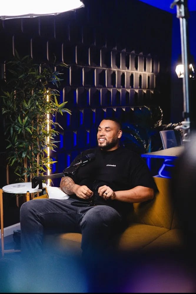 A UCP ambassador smiles while seated on a yellow couch in a modern setting with dark walls, a potted plant, and studio lighting.