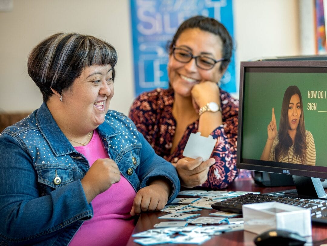 Two women are seated at a desk, smiling and looking at a computer screen showing a woman using sign language. One woman, holding photo prints, is excitedly discussing the benefits of adult day programs for fostering inclusive communication.