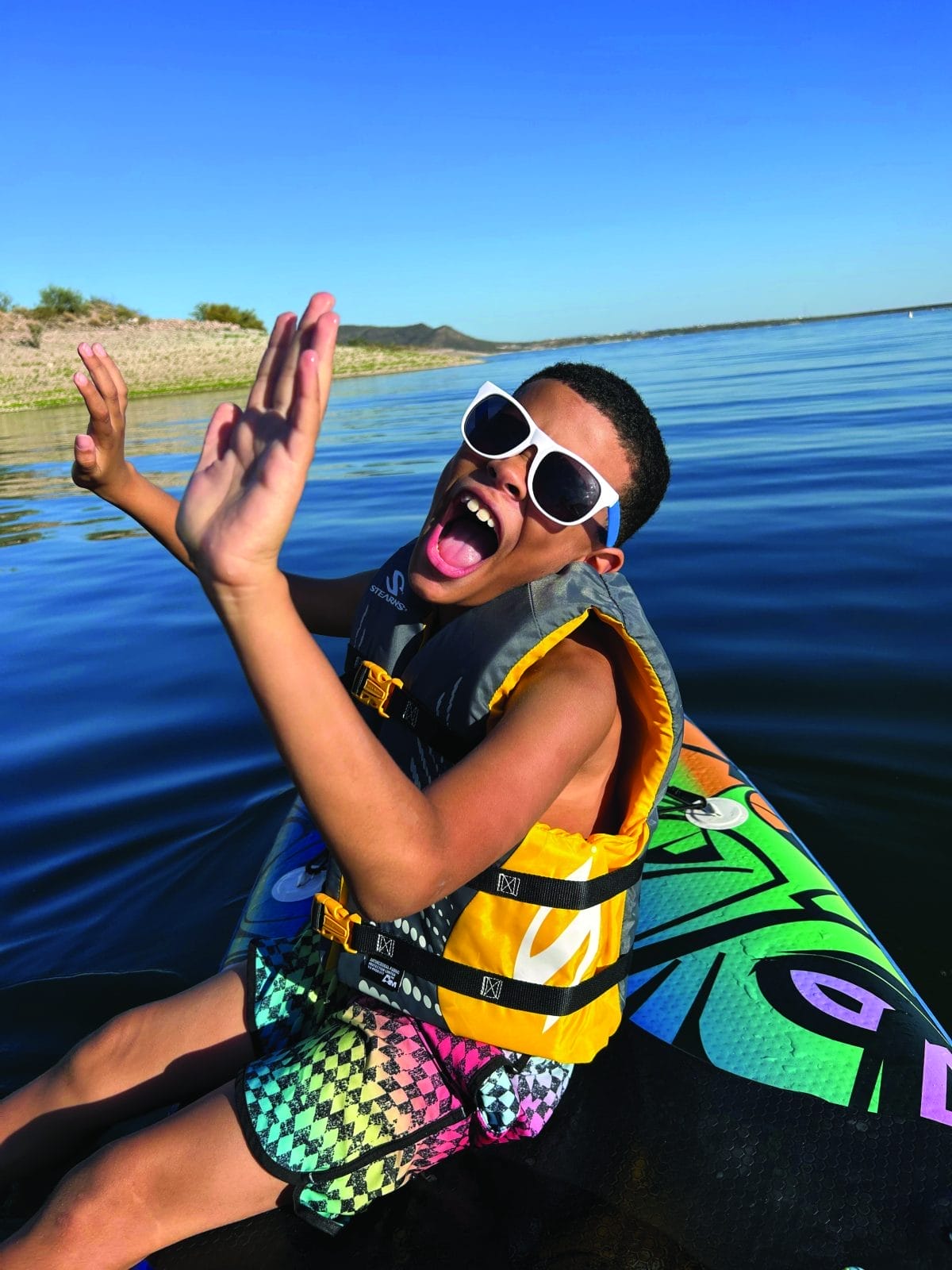 Nico RIdley in a life vest from UCP of Central AZ gives a high-five while sitting on a paddleboard on a lake.