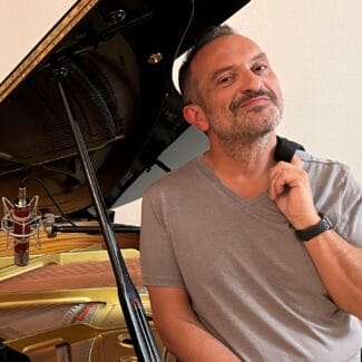 Carl Herrgesell smiling, leaning on a grand piano with its lid open, touching his chin thoughtfully, in a room with a beige wall, representing the united cerebral palsy of central Arizona.