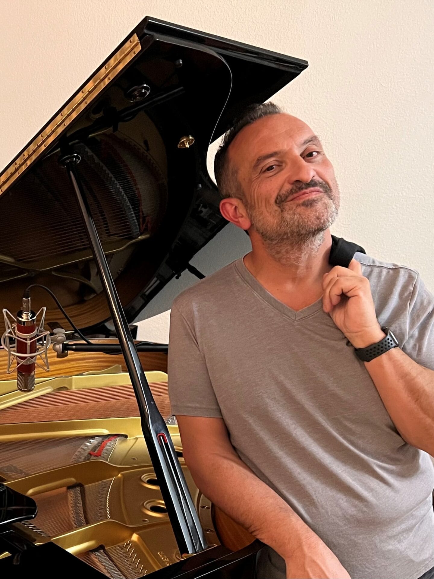 Carl Herrgesell smiling, leaning on a grand piano with its lid open, touching his chin thoughtfully, in a room with a beige wall, representing the united cerebral palsy of central Arizona.