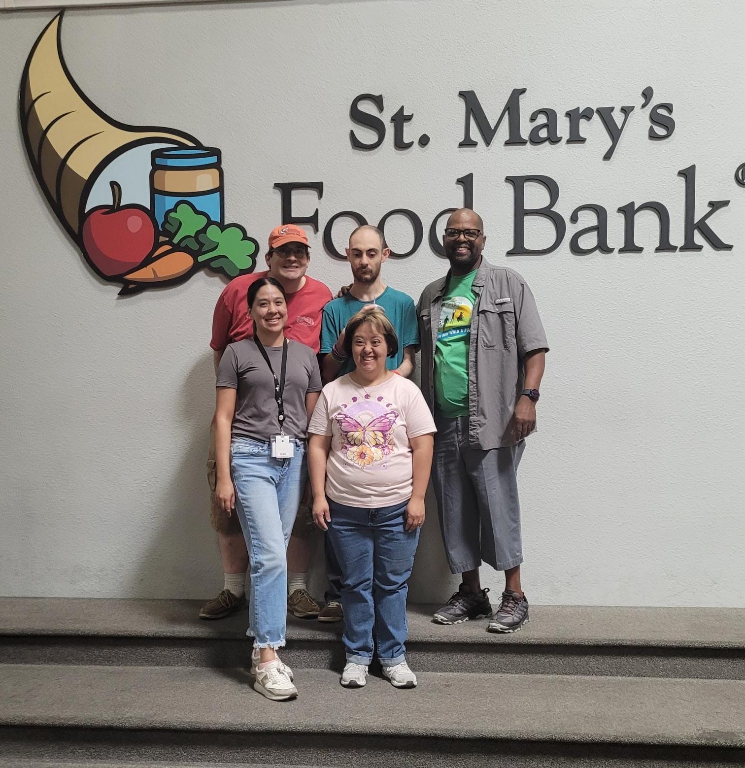 Five people stand in front of a wall with the logo and text "St. Mary's Food Bank." The group appears to be posing for a photo, with one member proudly displaying an AAC communication device.