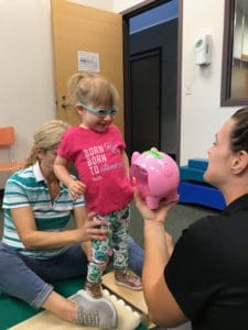 A little girl is engaged in pediatric therapy while playing with a piggy bank.