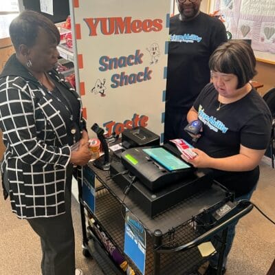 Two people assist a customer at a mobile snack cart labeled "YUMees Snack Shack." They are wearing matching black shirts, creating an inviting atmosphere similar to the community spirit found at adult day programs.