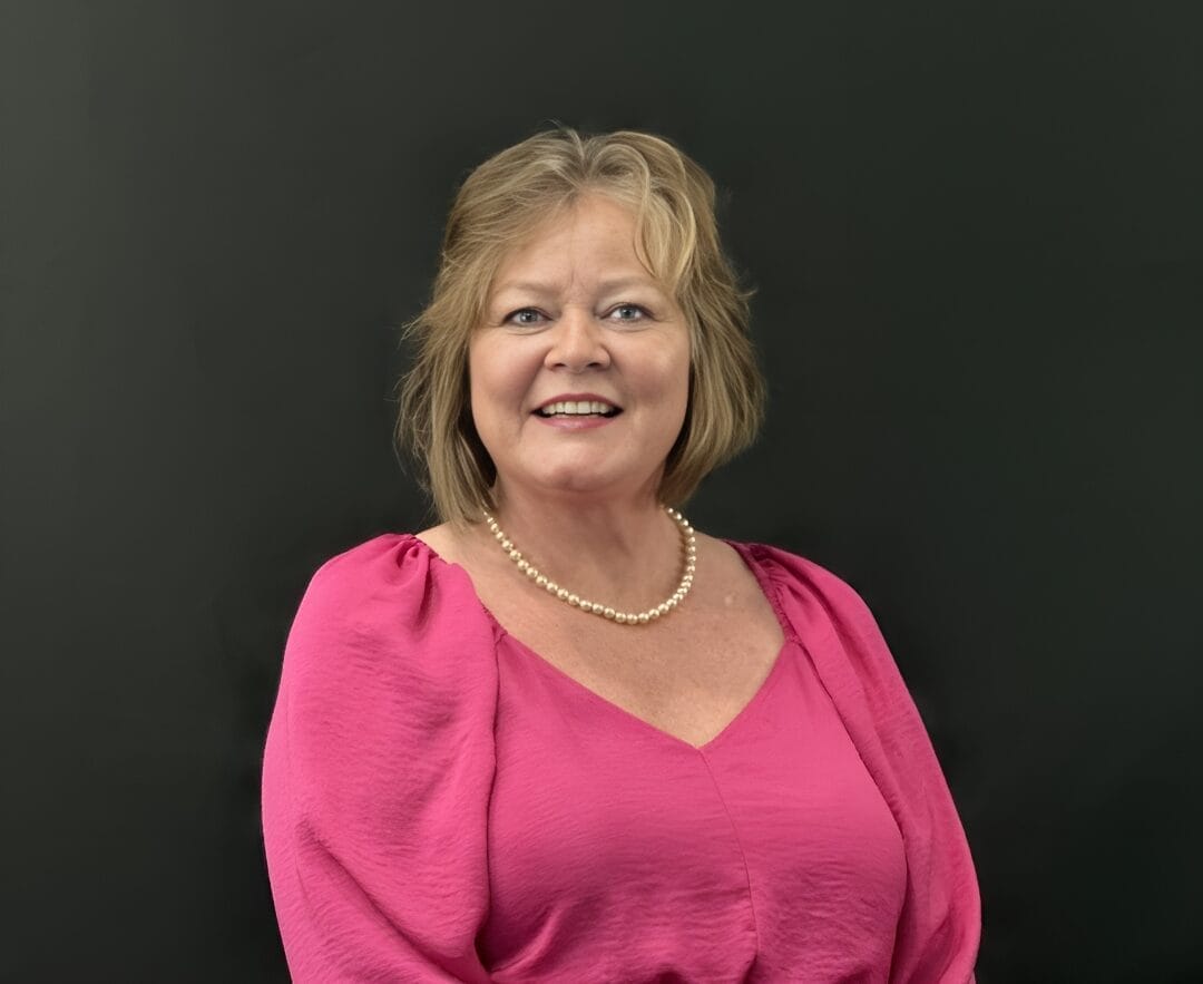 Woman with shoulder-length blonde hair, wearing a pink blouse and pearl necklace, smiling in front of a dark background. This engaging image could represent the welcoming environment found in adult day programs near me.