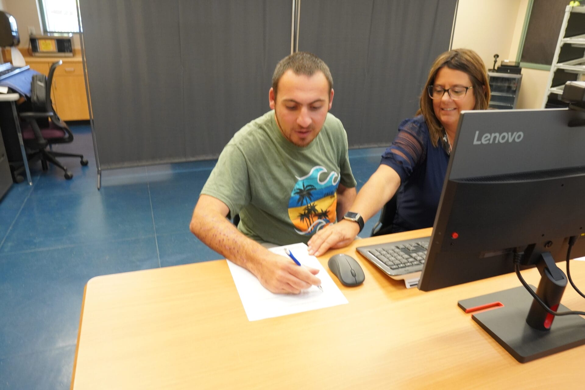 A person in a green T-shirt is sitting at a desk with a woman who is pointing at a piece of paper he is signing. A computer monitor, often used to browse adult day programs north Phoenix, is on the desk.