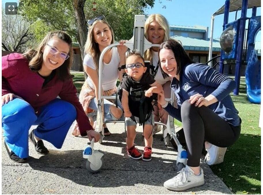 A young child using a walker is surrounded by four smiling adults from UCP of Central AZ, enjoying the sunny day near a playground.