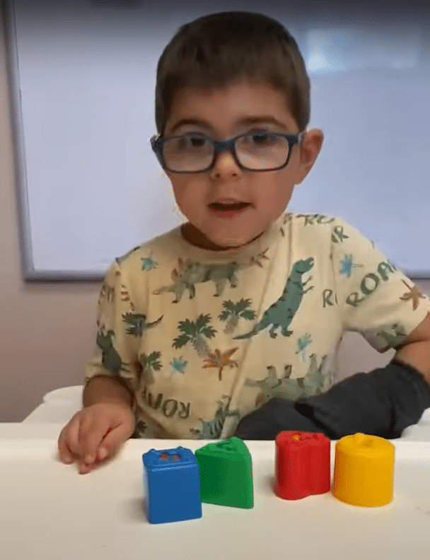 A young boy with glasses sitting at a table with colorful blocks in a home and community based services setting.