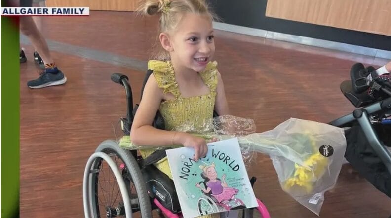 Norah smiling, holding her book titled "Moo Moo's World" at UCP of Central AZ.
