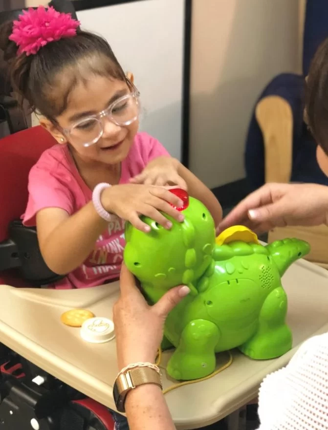 A young girl with cerebral palsy is playing with a toy dinosaur during a pediatric therapy session at home.