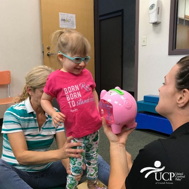 A young girl with cerebral palsy of Arizona is playing with a pink piggy bank at an early learning center.